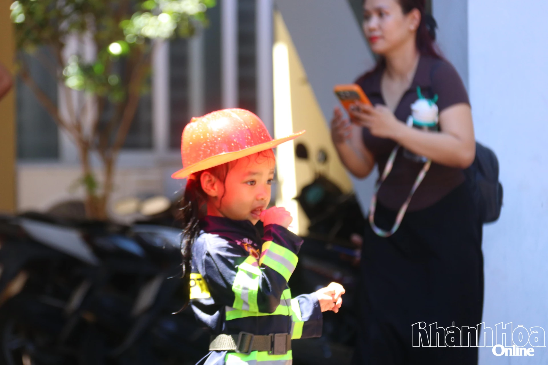 Một chiến sĩ nhí mặc đồng phục đang quan sát từ xa đồng đội thực hành chữa cháy bằng vòi phun nước.