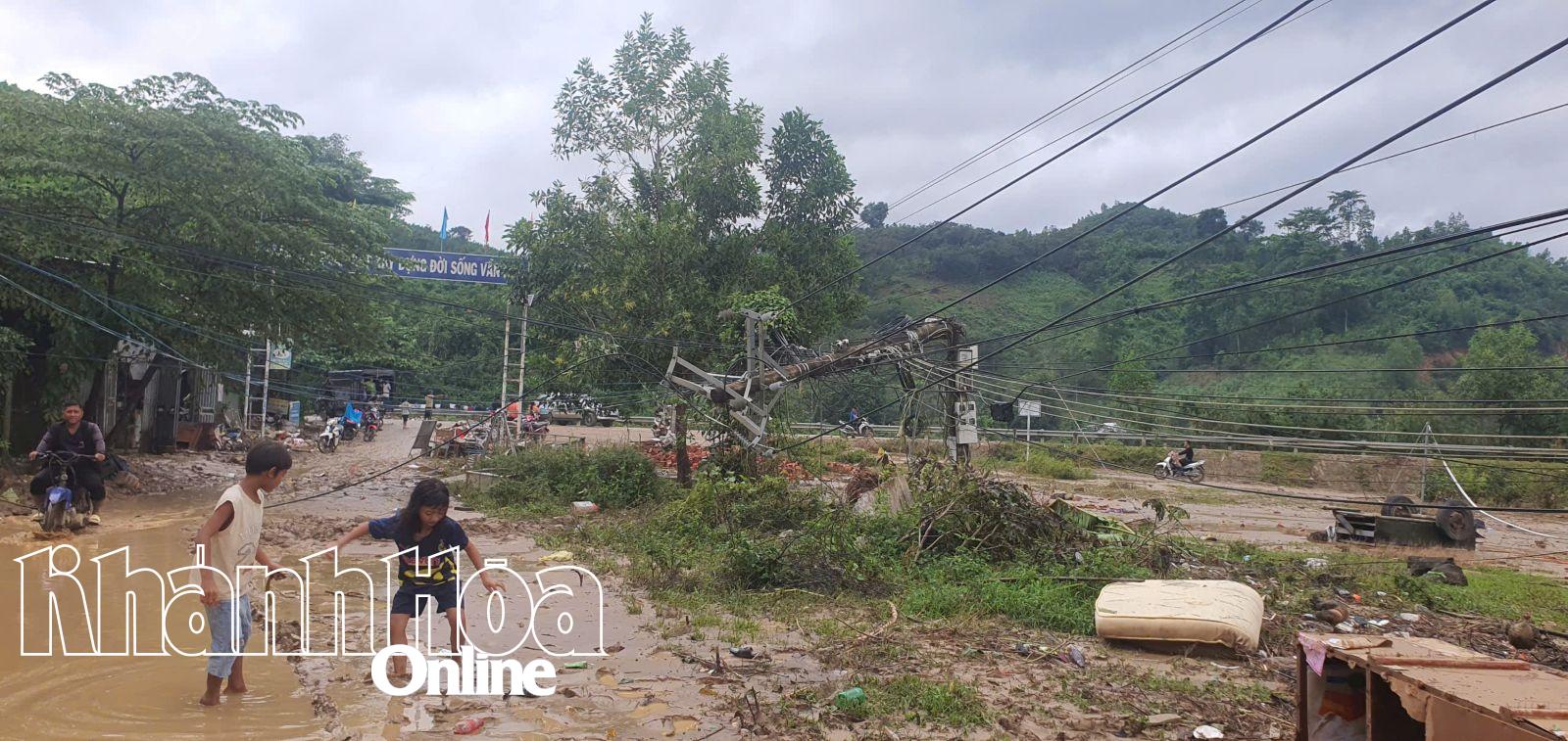 Trẻ em cẩn thận di chuyển qua cung đường đầy bùn đất, phía xa là một trụ điện đã bị gãy gục