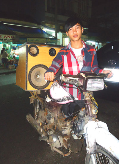 Một người hát rong bên chiếc xe gắn máy cũ và thùng đồ nghề.